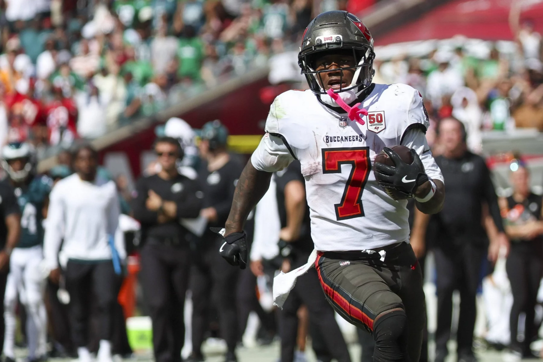 Bucky Irving - Ein Footballspieler der Tampa Bay Buccaneers, der die Trikotnummer 7 und schwarze Handschuhe trägt, läuft während eines Spiels gegen Seattle mit dem Ball. Unscharfe Gegner und Mannschaftskameraden füllen das Spielfeld, und auf den Sitzen im Stadion ist eine jubelnde Menge zu sehen. Diese Beschreibung wurde mit der FootballR KI automatisch generiert.