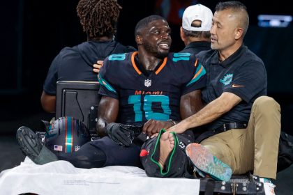 Tyreek Hill, der Dolphins-Star mit der Trikotnummer 10, sitzt ohne Schuh und mit bandagiertem linken Fuß auf einem Krankenwagen. Er wirkt emotional und wird von einem Mitarbeiter getröstet, als er vom Spielfeld gebracht wird, während andere Teammitglieder im Hintergrund zu sehen sind. Diese Beschreibung wurde mit der FootballR KI automatisch generiert.