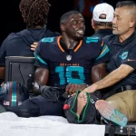 Tyreek Hill, der Dolphins-Star mit der Trikotnummer 10, sitzt ohne Schuh und mit bandagiertem linken Fuß auf einem Krankenwagen. Er wirkt emotional und wird von einem Mitarbeiter getröstet, als er vom Spielfeld gebracht wird, während andere Teammitglieder im Hintergrund zu sehen sind. Diese Beschreibung wurde mit der FootballR KI automatisch generiert.