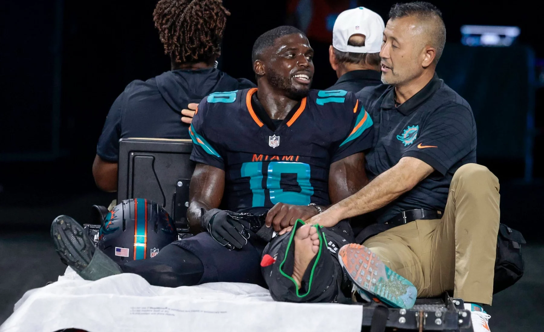 Tyreek Hill, der Dolphins-Star mit der Trikotnummer 10, sitzt ohne Schuh und mit bandagiertem linken Fuß auf einem Krankenwagen. Er wirkt emotional und wird von einem Mitarbeiter getröstet, als er vom Spielfeld gebracht wird, während andere Teammitglieder im Hintergrund zu sehen sind. Diese Beschreibung wurde mit der FootballR KI automatisch generiert.