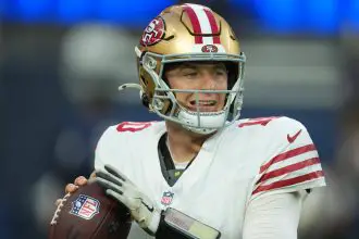 Ein Footballspieler im weißen Trikot der San Francisco 49ers und mit goldenem Helm, der Mac Jones ähnelt, hält einen Football und bereitet sich auf den Wurf vor. Das ikonische 49ers-Logo leuchtet auf seinem Helm, während er das Spielfeld abtastet. Die blauen Stadionsitze sind im Hintergrund verschwommen. Diese Beschreibung wurde mit der FootballR KI automatisch generiert.