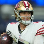 Ein Footballspieler im weißen Trikot der San Francisco 49ers und mit goldenem Helm, der Mac Jones ähnelt, hält einen Football und bereitet sich auf den Wurf vor. Das ikonische 49ers-Logo leuchtet auf seinem Helm, während er das Spielfeld abtastet. Die blauen Stadionsitze sind im Hintergrund verschwommen. Diese Beschreibung wurde mit der FootballR KI automatisch generiert.