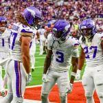 Drei Football-Spieler der Minnesota Vikings in weißen Trikots und lila Helmen feiern ausgelassen in der Endzone während des NFL International Series-Spiels der Vikings gegen die Browns in London, während die Zuschauer und andere Spieler den Stadionhintergrund füllen. Diese Beschreibung wurde mit der FootballR KI automatisch generiert.