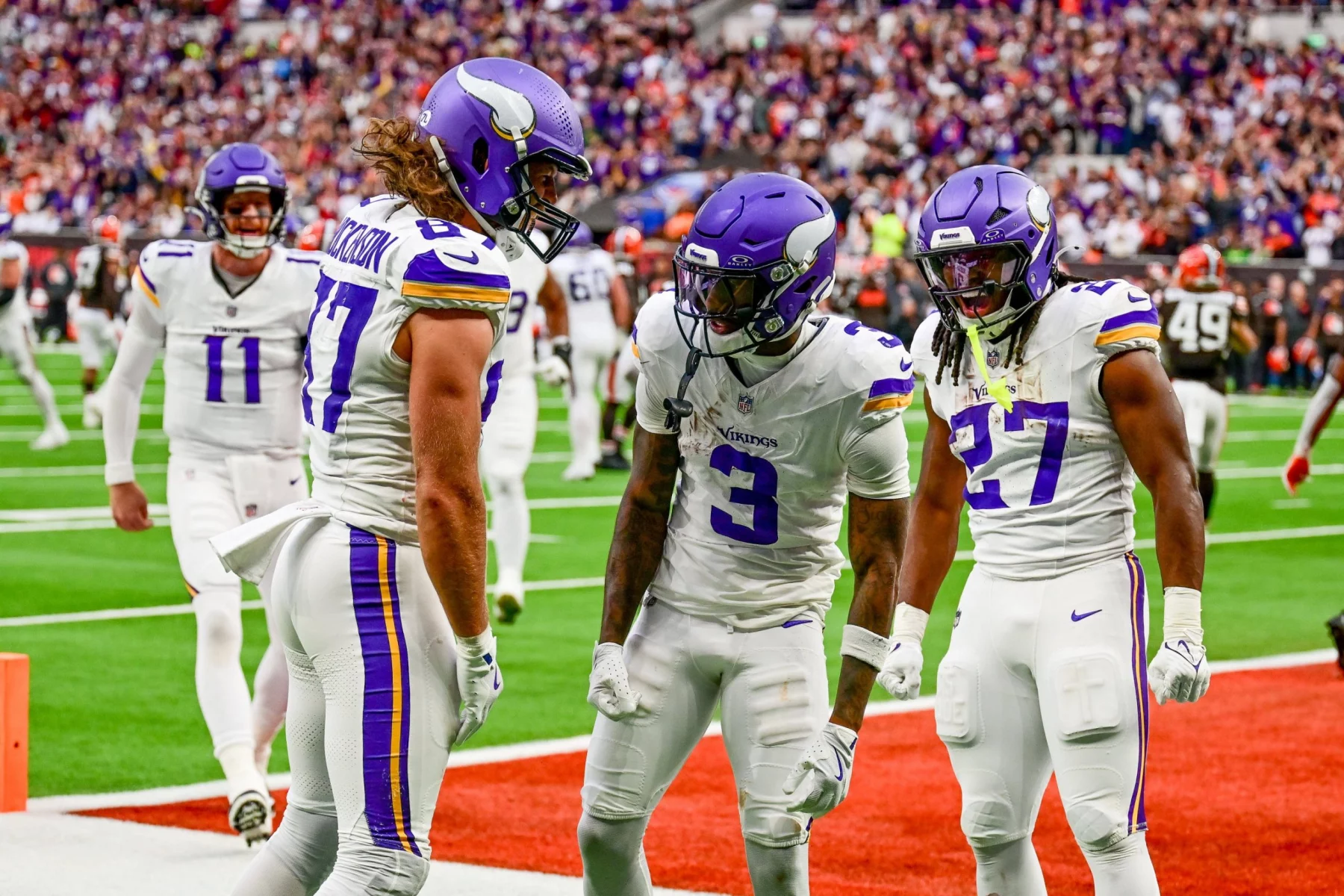 Drei Football-Spieler der Minnesota Vikings in weißen Trikots und lila Helmen feiern ausgelassen in der Endzone während des NFL International Series-Spiels der Vikings gegen die Browns in London, während die Zuschauer und andere Spieler den Stadionhintergrund füllen. Diese Beschreibung wurde mit der FootballR KI automatisch generiert.