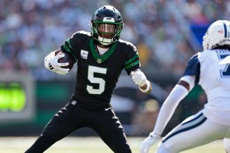 Star-Receiver Garrett Wilson von den Jets, der ein schwarzes Trikot mit der Nummer 5 trägt, sprintet mit Ball und Visier auf einen Gegner in Weiß und Blau zu, während im Hintergrund ein verschwommenes Stadion voller Fans zu sehen ist. Diese Beschreibung wurde mit der FootballR KI automatisch generiert.