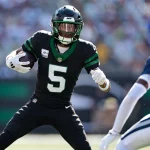 Star-Receiver Garrett Wilson von den Jets, der ein schwarzes Trikot mit der Nummer 5 trägt, sprintet mit Ball und Visier auf einen Gegner in Weiß und Blau zu, während im Hintergrund ein verschwommenes Stadion voller Fans zu sehen ist. Diese Beschreibung wurde mit der FootballR KI automatisch generiert.