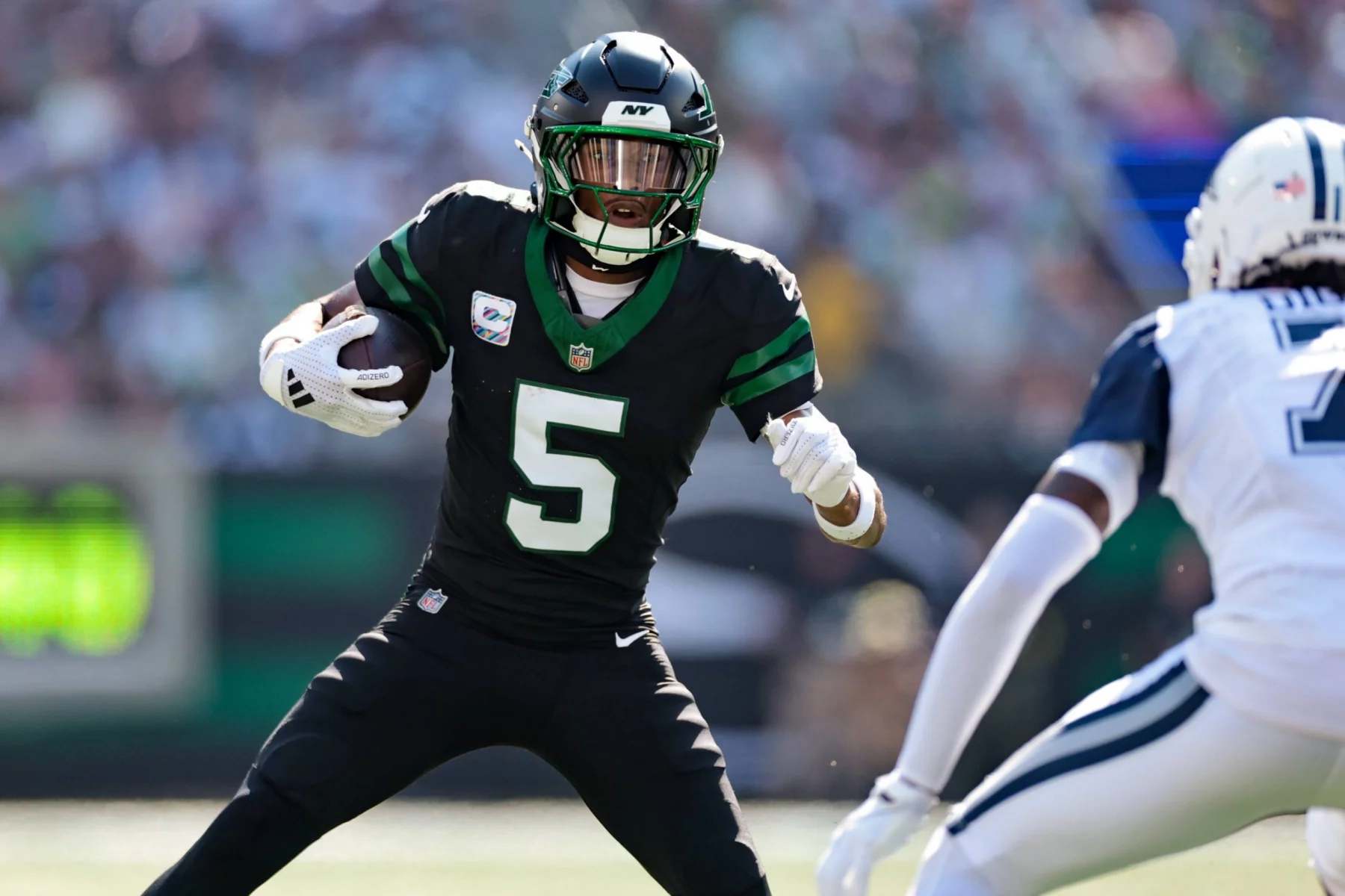 Star-Receiver Garrett Wilson von den Jets, der ein schwarzes Trikot mit der Nummer 5 trägt, sprintet mit Ball und Visier auf einen Gegner in Weiß und Blau zu, während im Hintergrund ein verschwommenes Stadion voller Fans zu sehen ist. Diese Beschreibung wurde mit der FootballR KI automatisch generiert.