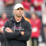 Ein Mann mit weißer Mütze und schwarzem "Arizona Cardinals"-Kapuzenpulli steht mit verschränkten Armen da und wirkt nachdenklich. Cardinals-Coach Jonathan Gannon steht auf einem Football-Feld mit unscharfen Menschen in roten Hemden und Stadionsitzen im Hintergrund. Diese Beschreibung wurde mit der FootballR KI automatisch generiert.