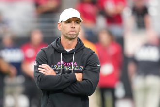 Ein Mann mit weißer Mütze und schwarzem "Arizona Cardinals"-Kapuzenpulli steht mit verschränkten Armen da und wirkt nachdenklich. Cardinals-Coach Jonathan Gannon steht auf einem Football-Feld mit unscharfen Menschen in roten Hemden und Stadionsitzen im Hintergrund. Diese Beschreibung wurde mit der FootballR KI automatisch generiert.