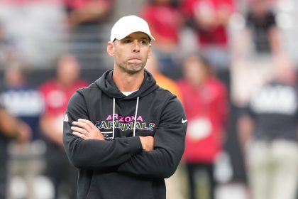 Ein Mann mit weißer Mütze und schwarzem "Arizona Cardinals"-Kapuzenpulli steht mit verschränkten Armen da und wirkt nachdenklich. Cardinals-Coach Jonathan Gannon steht auf einem Football-Feld mit unscharfen Menschen in roten Hemden und Stadionsitzen im Hintergrund. Diese Beschreibung wurde mit der FootballR KI automatisch generiert.