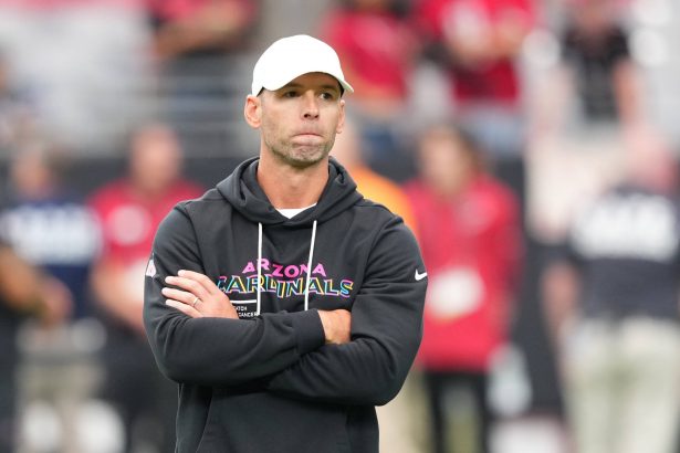 Ein Mann mit weißer Mütze und schwarzem "Arizona Cardinals"-Kapuzenpulli steht mit verschränkten Armen da und wirkt nachdenklich. Cardinals-Coach Jonathan Gannon steht auf einem Football-Feld mit unscharfen Menschen in roten Hemden und Stadionsitzen im Hintergrund. Diese Beschreibung wurde mit der FootballR KI automatisch generiert.