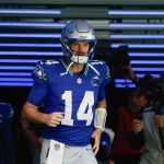 NFL Week 6 Prediction - Sam Darnold - Ein Football-Spieler in einem blauen Seattle Seahawks-Trikot mit der Nummer 14 und einem silbernen Helm läuft aus einem mit blauen Lichtern gesäumten Tunnel auf das Spielfeld und bringt Spannung und Vorfreude wie NFL Week 6 Predictions mit sich. Ein Kameramann filmt ihn, während ein anderer Spieler hinterherläuft. Diese Beschreibung wurde mit der FootballR KI automatisch generiert.