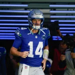 Week 9 - Divisional Round - Super Bowl LX - NFL Week 6 Prediction - Sam Darnold - Ein Football-Spieler in einem blauen Seattle Seahawks-Trikot mit der Nummer 14 und einem silbernen Helm läuft aus einem mit blauen Lichtern gesäumten Tunnel auf das Spielfeld und bringt Spannung und Vorfreude wie NFL Week 6 Predictions mit sich. Ein Kameramann filmt ihn, während ein anderer Spieler hinterherläuft. Diese Beschreibung wurde mit der FootballR KI automatisch generiert.