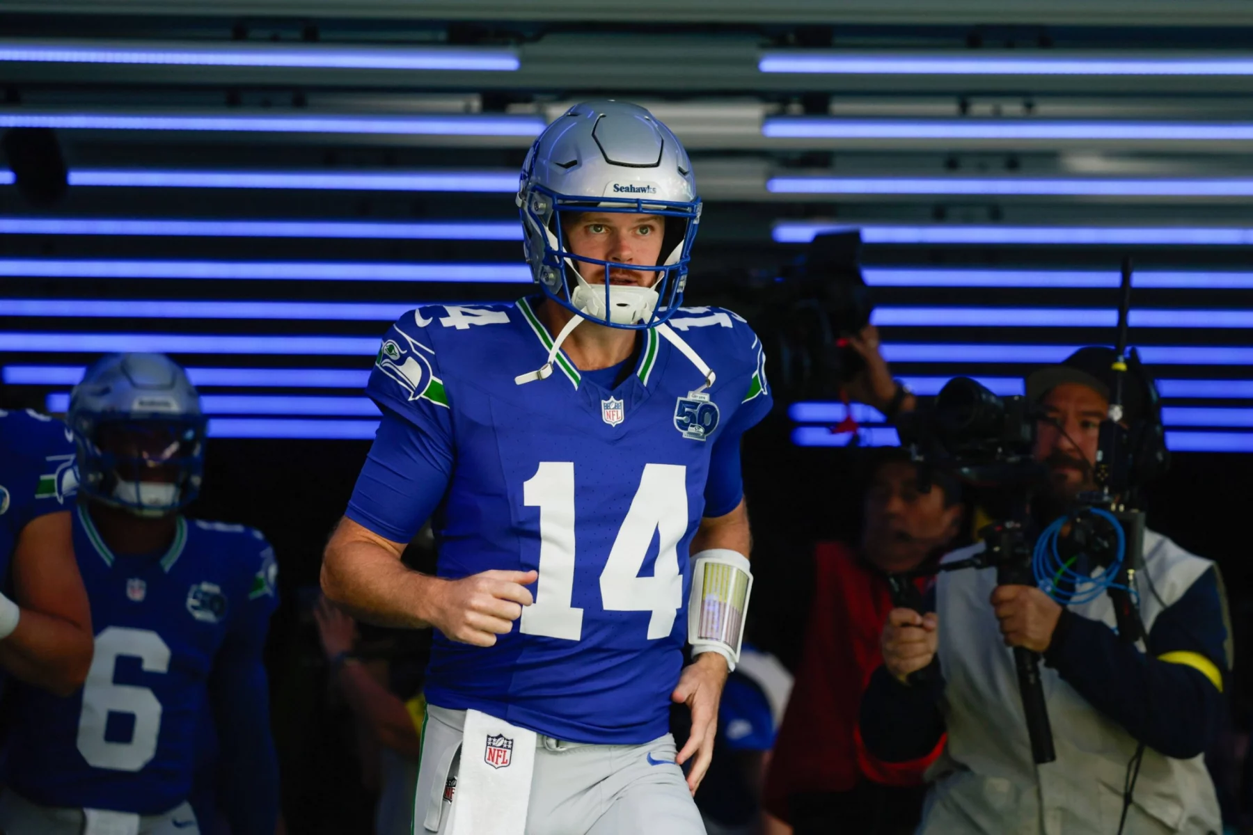 Week 9 - Divisional Round - Super Bowl LX - NFL Week 6 Prediction - Sam Darnold - Ein Football-Spieler in einem blauen Seattle Seahawks-Trikot mit der Nummer 14 und einem silbernen Helm läuft aus einem mit blauen Lichtern gesäumten Tunnel auf das Spielfeld und bringt Spannung und Vorfreude wie NFL Week 6 Predictions mit sich. Ein Kameramann filmt ihn, während ein anderer Spieler hinterherläuft. Diese Beschreibung wurde mit der FootballR KI automatisch generiert.