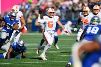 BucsQuarterback Baker Mayfield (#6) in einem weiß-orangenen Trikot läuft nach rechts und bereitet sich auf den Wurf vor, während die Verteidiger in der NFL Woche 5 näher kommen. Seine Teamkollegen schützen ihn auf einem sonnigen Feld, mit einer verschwommenen Menschenmenge im Hintergrund. Diese Beschreibung wurde mit der FootballR KI automatisch generiert.
