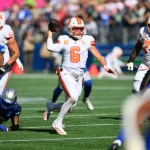 BucsQuarterback Baker Mayfield (#6) in einem weiß-orangenen Trikot läuft nach rechts und bereitet sich auf den Wurf vor, während die Verteidiger in der NFL Woche 5 näher kommen. Seine Teamkollegen schützen ihn auf einem sonnigen Feld, mit einer verschwommenen Menschenmenge im Hintergrund. Diese Beschreibung wurde mit der FootballR KI automatisch generiert.