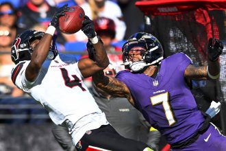 Ein Spieler der Houston Texans in einem weißen Trikot versucht, einen Football zu fangen, während sich die Offense der Baltimore Ravens ausstreckt, um ihn zu blocken. Beide Athleten sind in Bewegung und konzentrieren sich auf den Ball. Der Stadionhintergrund ist unscharf und die rote Ausrüstung sichtbar. Diese Beschreibung wurde mit der FootballR KI automatisch generiert.
