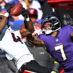 Ein Spieler der Houston Texans in einem weißen Trikot versucht, einen Football zu fangen, während sich die Offense der Baltimore Ravens ausstreckt, um ihn zu blocken. Beide Athleten sind in Bewegung und konzentrieren sich auf den Ball. Der Stadionhintergrund ist unscharf und die rote Ausrüstung sichtbar. Diese Beschreibung wurde mit der FootballR KI automatisch generiert.