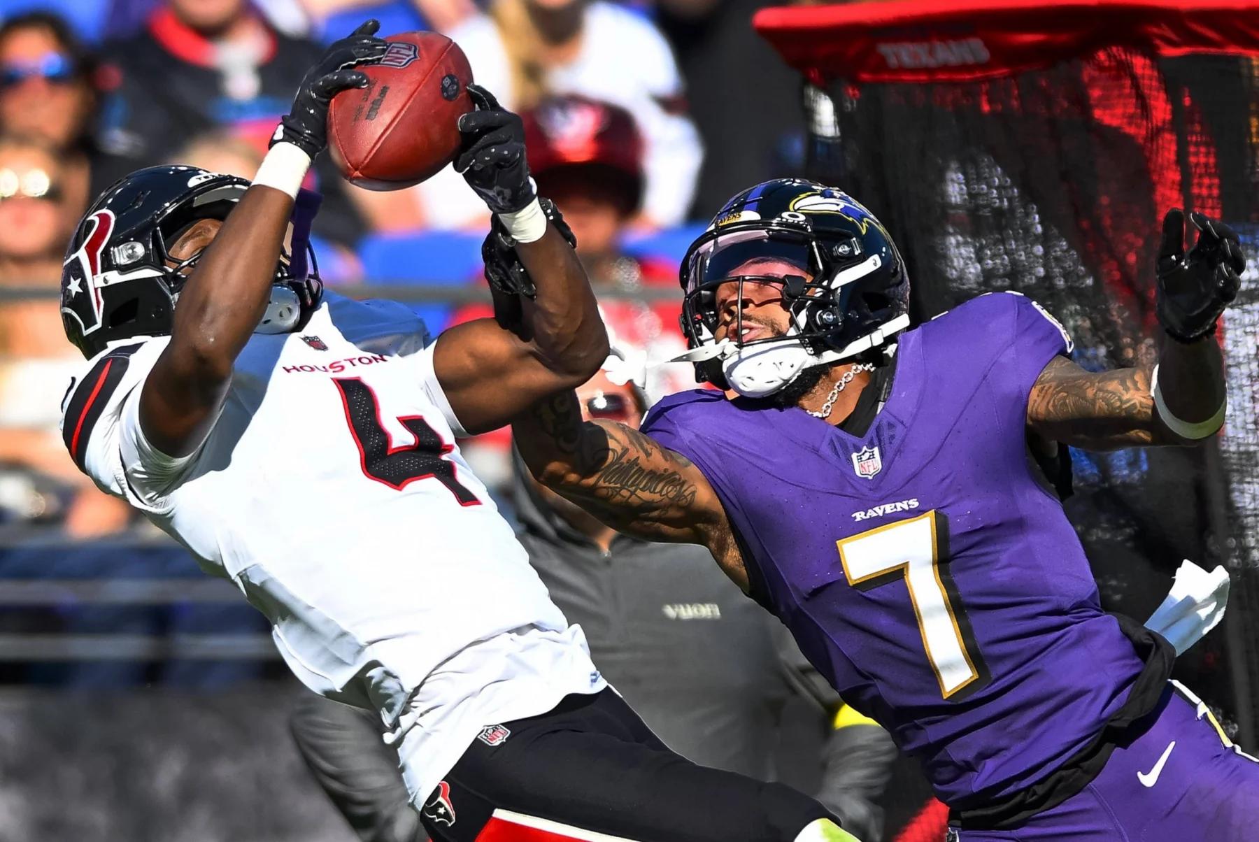 Ein Spieler der Houston Texans in einem weißen Trikot versucht, einen Football zu fangen, während sich die Offense der Baltimore Ravens ausstreckt, um ihn zu blocken. Beide Athleten sind in Bewegung und konzentrieren sich auf den Ball. Der Stadionhintergrund ist unscharf und die rote Ausrüstung sichtbar. Diese Beschreibung wurde mit der FootballR KI automatisch generiert.