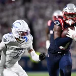 Ein Footballspieler der New England Patriots - Stefon Diggs - in einer Marineuniform läuft mit dem Ball, dicht verfolgt von einem Verteidiger der Buffalo Bills während eines NFL-Spiels. Der unscharfe Hintergrund hebt die Aktion hervor, als die Patriots gegen Stefon Diggs und die Bills antreten. Diese Beschreibung wurde mit der FootballR KI automatisch generiert.