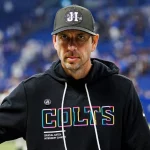 Ein Mann, der dem Colts-Trainer Shane Steichen ähnelt, steht in einem Stadion und trägt einen schwarzen "Colts"-Kapuzenpullover mit bunten Logos. Der Hintergrund ist unscharf mit blauen Stadionsitzen und Menschen, während er ernst in die Kamera blickt. Diese Beschreibung wurde mit der FootballR KI automatisch generiert.