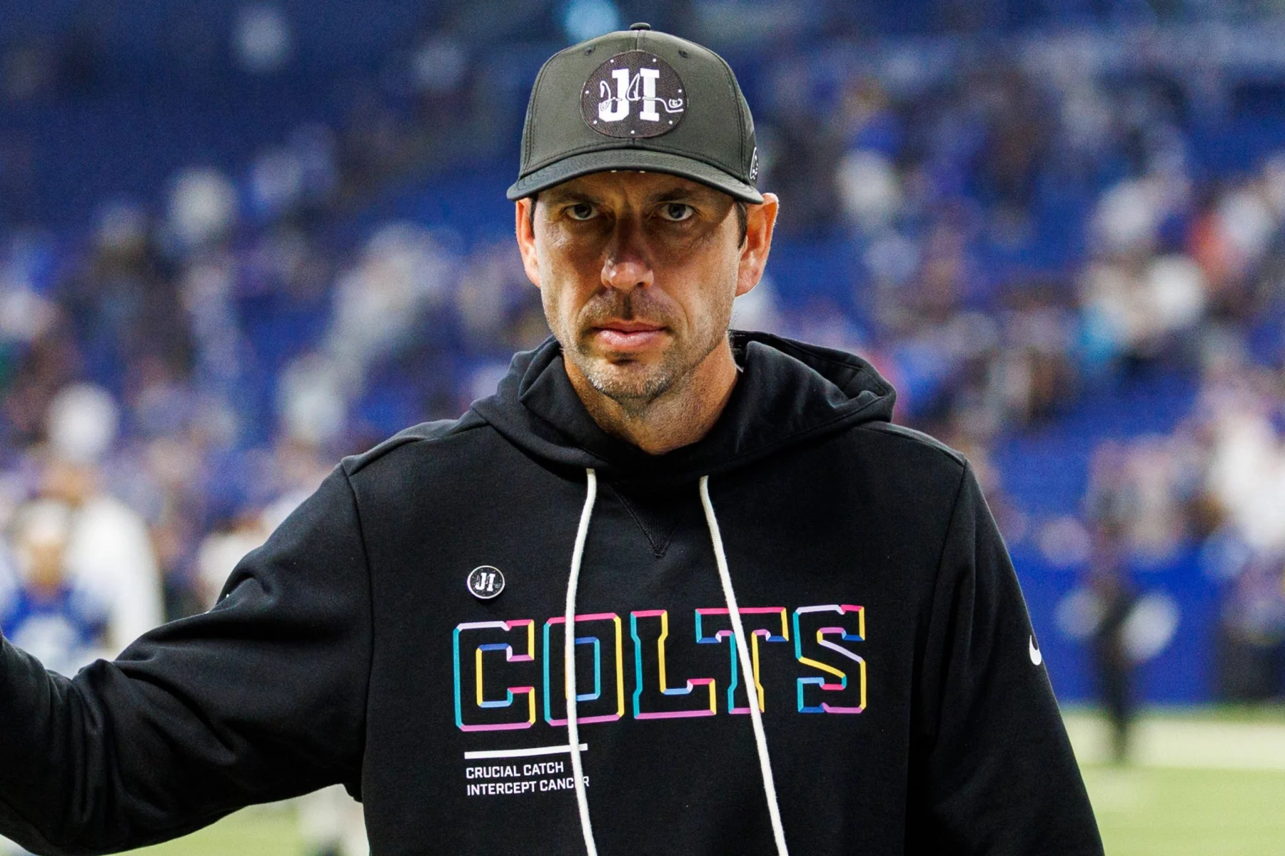 Ein Mann, der dem Colts-Trainer Shane Steichen ähnelt, steht in einem Stadion und trägt einen schwarzen "Colts"-Kapuzenpullover mit bunten Logos. Der Hintergrund ist unscharf mit blauen Stadionsitzen und Menschen, während er ernst in die Kamera blickt. Diese Beschreibung wurde mit der FootballR KI automatisch generiert.