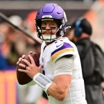 Ein Football-Spieler in einer weißen Vikings-Uniform und einem lila Helm hält einen Football und bereitet sich auf den Wurf vor. Er blickt konzentriert auf das Spielfeld und erinnert an Carson Wentz. Der unscharfe Hintergrund unterstreicht die Atmosphäre des Spiels mit dem Personal an der Seitenlinie und einer lebhaften Menge. Diese Beschreibung wurde mit der FootballR KI automatisch generiert.