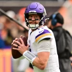 Ein Football-Spieler in einer weißen Vikings-Uniform und einem lila Helm hält einen Football und bereitet sich auf den Wurf vor. Er blickt konzentriert auf das Spielfeld und erinnert an Carson Wentz. Der unscharfe Hintergrund unterstreicht die Atmosphäre des Spiels mit dem Personal an der Seitenlinie und einer lebhaften Menge. Diese Beschreibung wurde mit der FootballR KI automatisch generiert.