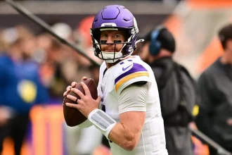 Ein Football-Spieler in einer weißen Vikings-Uniform und einem lila Helm hält einen Football und bereitet sich auf den Wurf vor. Er blickt konzentriert auf das Spielfeld und erinnert an Carson Wentz. Der unscharfe Hintergrund unterstreicht die Atmosphäre des Spiels mit dem Personal an der Seitenlinie und einer lebhaften Menge. Diese Beschreibung wurde mit der FootballR KI automatisch generiert.