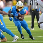Ein Footballspieler im blauen Trikot der Los Angeles Chargers läuft mit dem Ball und weicht Verteidigern in Weiß und Rot aus. Teamkollegen blockieren, während Runningback Omarion Hampton auf dem grünen Feld nach vorne stürmt, das Gesicht hinter einer gelben Gesichtsmaske. Diese Beschreibung wurde mit der FootballR KI automatisch generiert.