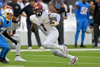 Deebo Samuel, Commanders, in einer weißen Uniform rennt mit dem Ball, verfolgt von einem Spieler der Los Angeles Chargers in Blau. Die actiongeladene Szene, die an den dynamischen Stil von Deebo Samuel erinnert, spielt sich während eines Spiels ab, mit Spielern und Mitarbeitern im Hintergrund. Diese Beschreibung wurde mit der FootballR KI automatisch generiert.