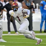 Deebo Samuel, Commanders, in einer weißen Uniform rennt mit dem Ball, verfolgt von einem Spieler der Los Angeles Chargers in Blau. Die actiongeladene Szene, die an den dynamischen Stil von Deebo Samuel erinnert, spielt sich während eines Spiels ab, mit Spielern und Mitarbeitern im Hintergrund. Diese Beschreibung wurde mit der FootballR KI automatisch generiert.