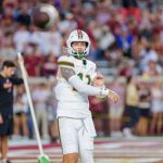Carson Beck - Ein College-Football-Spieler der Woche im weißen Trikot der Miami Hurricanes, Nummer 12, bereitet sich darauf vor, einen Football in der Luft zu fangen. Er trägt einen Helm und eine Armmanschette, während das verschwommene Stadionpublikum, einige in kastanienbraun, während der Woche 6 zusieht. Diese Beschreibung wurde mit der FootballR KI automatisch generiert.