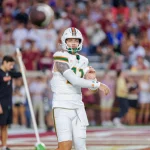 Carson Beck - Ein College-Football-Spieler der Woche im weißen Trikot der Miami Hurricanes, Nummer 12, bereitet sich darauf vor, einen Football in der Luft zu fangen. Er trägt einen Helm und eine Armmanschette, während das verschwommene Stadionpublikum, einige in kastanienbraun, während der Woche 6 zusieht. Diese Beschreibung wurde mit der FootballR KI automatisch generiert.