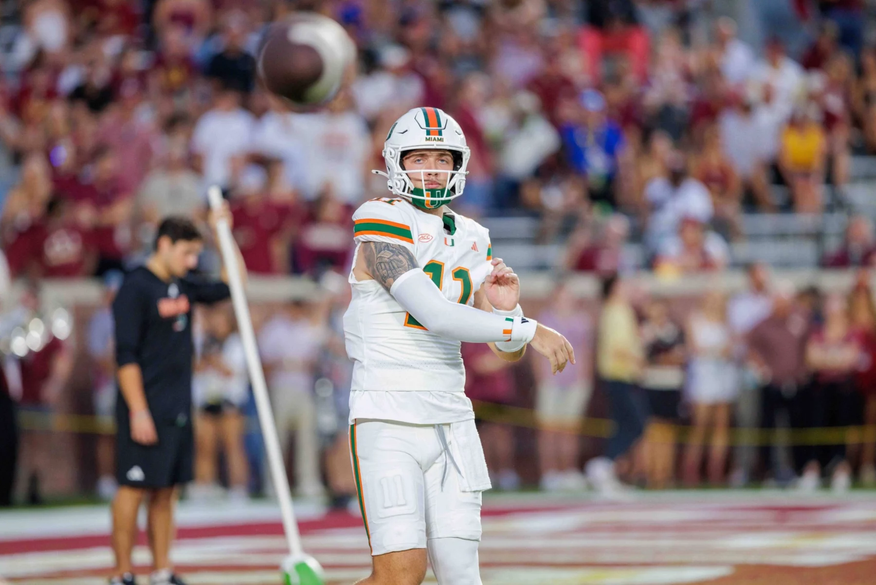 Carson Beck - Ein College-Football-Spieler der Woche im weißen Trikot der Miami Hurricanes, Nummer 12, bereitet sich darauf vor, einen Football in der Luft zu fangen. Er trägt einen Helm und eine Armmanschette, während das verschwommene Stadionpublikum, einige in kastanienbraun, während der Woche 6 zusieht. Diese Beschreibung wurde mit der FootballR KI automatisch generiert.
