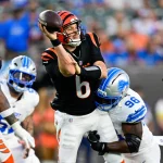 Der Quarterback der Cincinnati Bengals, Jake Browning, in einer schwarzen Uniform mit orangefarbenen Streifen bereitet sich darauf vor, einen Football zu werfen, während er von einem Verteidiger der Detroit Lions während eines intensiven Spiels angegangen wird. Der Coach sieht zu und denkt über einen möglichen QB-Wechsel nach. Diese Beschreibung wurde mit der FootballR KI automatisch generiert.