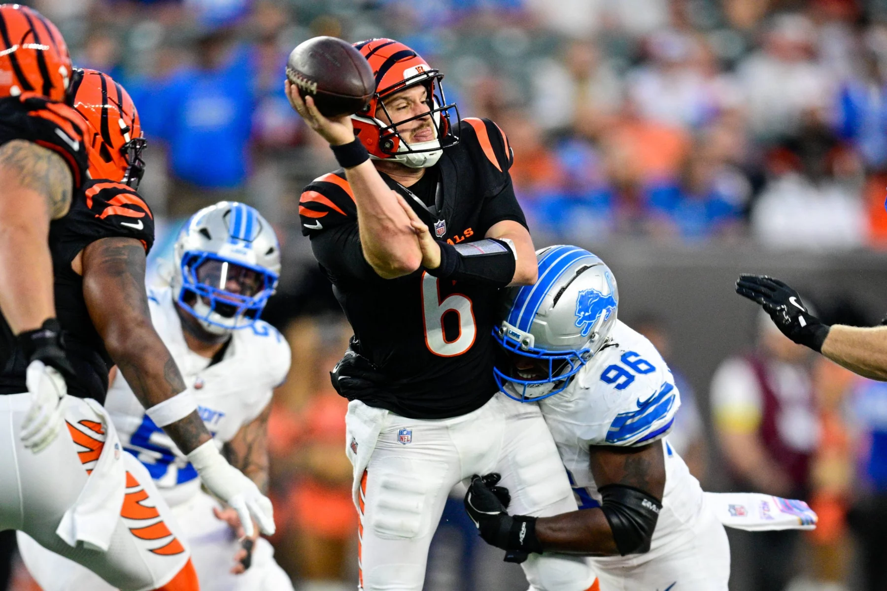 Der Quarterback der Cincinnati Bengals, Jake Browning, in einer schwarzen Uniform mit orangefarbenen Streifen bereitet sich darauf vor, einen Football zu werfen, während er von einem Verteidiger der Detroit Lions während eines intensiven Spiels angegangen wird. Der Coach sieht zu und denkt über einen möglichen QB-Wechsel nach. Diese Beschreibung wurde mit der FootballR KI automatisch generiert.