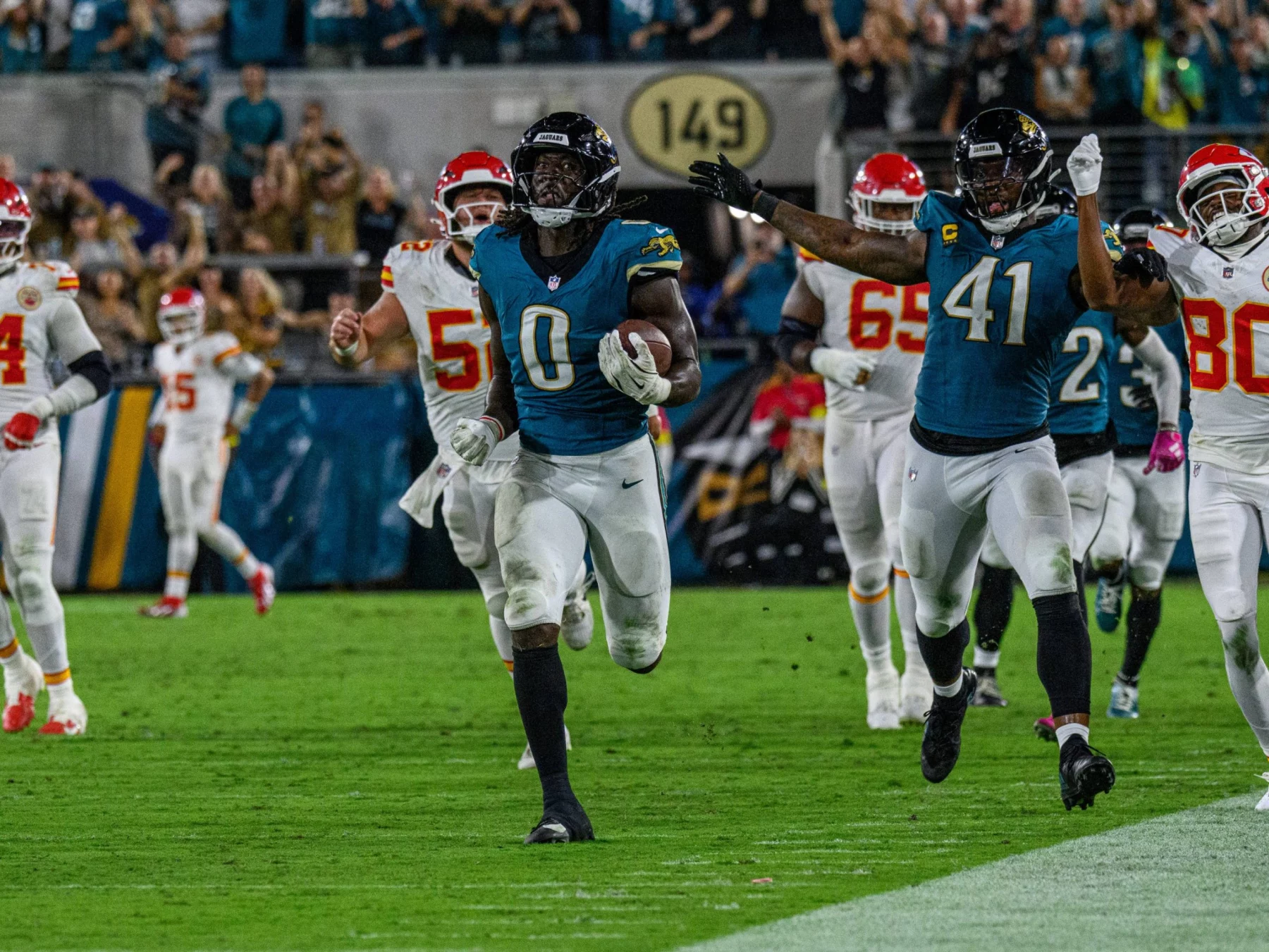 Ein Jacksonville Jaguars Spieler - Devin Lloyd - der Woche rennt mit dem Football in Richtung Endzone, dicht gefolgt von einem Teamkollegen und mehreren Verteidigern der Kansas City Chiefs in diesem spannenden NFL Woche 5 Spiel, während die Menge im Stadion jubelt. Diese Beschreibung wurde mit der FootballR KI automatisch generiert.