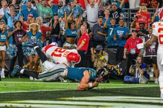 Ein Football-Spieler im schwarz-weißen Trikot der Jaguars, möglicherweise Trevor Lawrence, springt in die Endzone, während ein weiß-roter Verteidiger der Chiefs ihn angreift, während Cheerleader und Fans den dramatischen Moment auf der Torlinie lautstark bejubeln. Diese Beschreibung wurde mit der FootballR KI automatisch generiert.