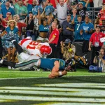 Ein Football-Spieler im schwarz-weißen Trikot der Jaguars, möglicherweise Trevor Lawrence, springt in die Endzone, während ein weiß-roter Verteidiger der Chiefs ihn angreift, während Cheerleader und Fans den dramatischen Moment auf der Torlinie lautstark bejubeln. Diese Beschreibung wurde mit der FootballR KI automatisch generiert.