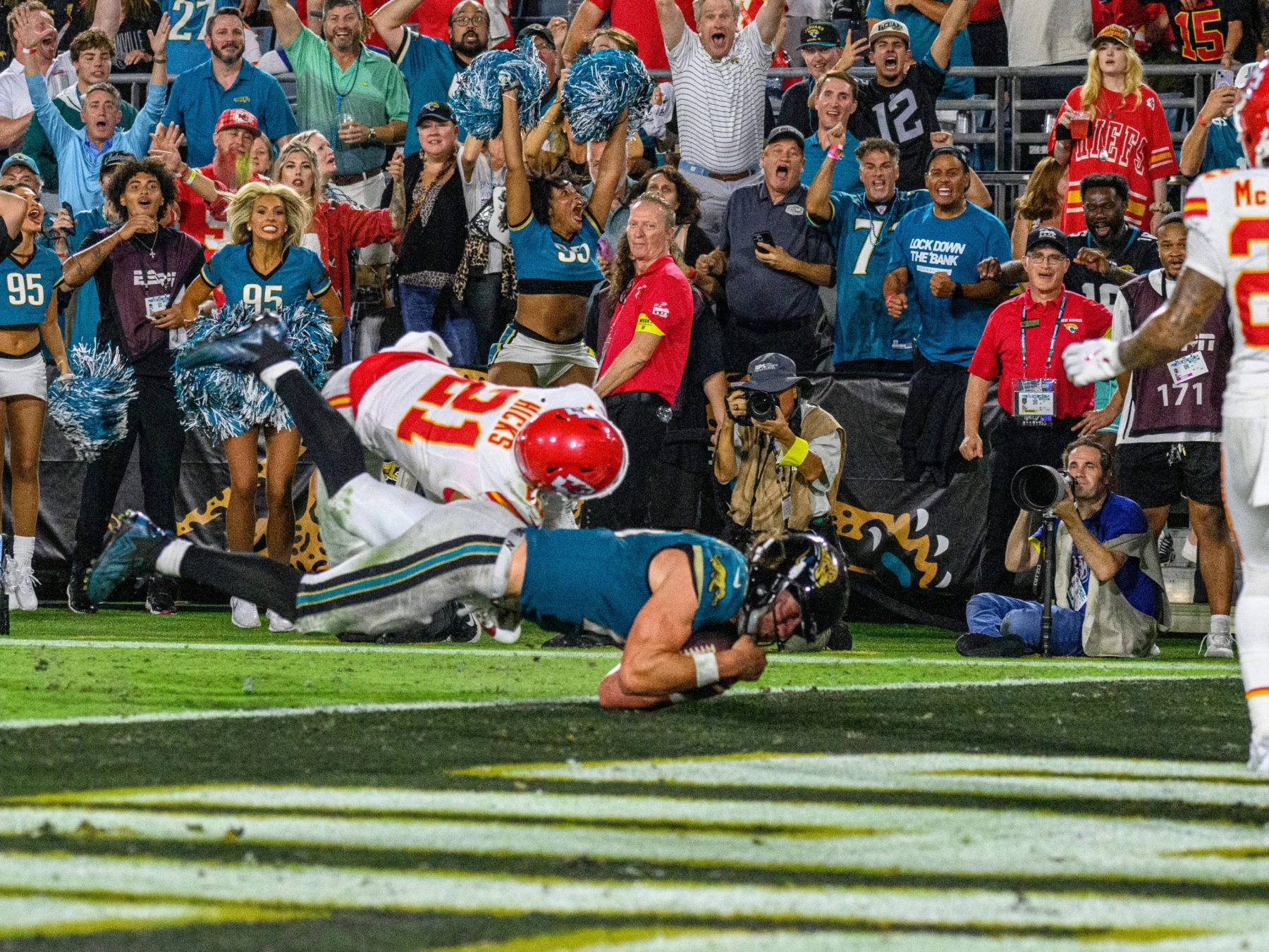 Ein Football-Spieler im schwarz-weißen Trikot der Jaguars, möglicherweise Trevor Lawrence, springt in die Endzone, während ein weiß-roter Verteidiger der Chiefs ihn angreift, während Cheerleader und Fans den dramatischen Moment auf der Torlinie lautstark bejubeln. Diese Beschreibung wurde mit der FootballR KI automatisch generiert.