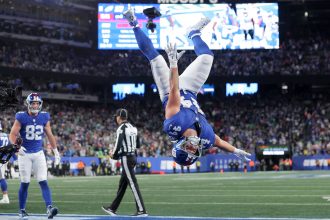 Ein Footballspieler der New York Giants, möglicherweise einer der Giants-Rookies, vollführt während eines Sensationssieges einen Rückwärtssalto in der Endzone, während jubelnde Fans und eine leuchtende Anzeigetafel das vollbesetzte Stadion erhellen. Diese Beschreibung wurde mit der FootballR KI automatisch generiert.