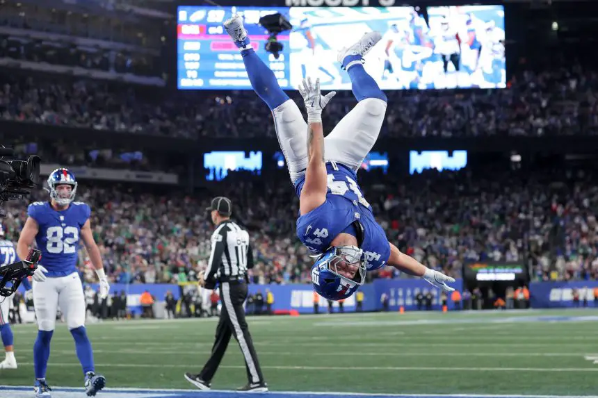 Ein Footballspieler der New York Giants, möglicherweise einer der Giants-Rookies, vollführt während eines Sensationssieges einen Rückwärtssalto in der Endzone, während jubelnde Fans und eine leuchtende Anzeigetafel das vollbesetzte Stadion erhellen. Diese Beschreibung wurde mit der FootballR KI automatisch generiert.
