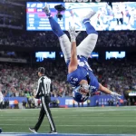 Ein Footballspieler der New York Giants, möglicherweise einer der Giants-Rookies, vollführt während eines Sensationssieges einen Rückwärtssalto in der Endzone, während jubelnde Fans und eine leuchtende Anzeigetafel das vollbesetzte Stadion erhellen. Diese Beschreibung wurde mit der FootballR KI automatisch generiert.