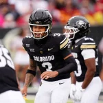 Kaidon Salter - In Week 7 des College Footballs ruft ein Spieler der Colorado Buffaloes, Nummer 3, im schwarzen Trikot ein Play aus. Umgeben von Teamkollegen, darunter Nummer 85, hebt er sich vor der unscharfen Kulisse der jubelnden Fans hervor. Diese Beschreibung wurde mit der FootballR KI automatisch generiert.