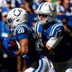 Zwei Footballspieler der Indianapolis Colts in blauen Uniformen und weißen Helmen spielen während eines Spiels. Der Quarterback Daniel Jones scheint den Ball an Jonathan Taylor abzugeben. Eine blau-weiß gekleidete Menschenmenge ist im sonnenbeschienenen Hintergrund verschwommen. Diese Beschreibung wurde mit der FootballR KI automatisch generiert.
