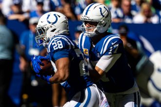 Zwei Footballspieler der Indianapolis Colts in blauen Uniformen und weißen Helmen spielen während eines Spiels. Der Quarterback Daniel Jones scheint den Ball an Jonathan Taylor abzugeben. Eine blau-weiß gekleidete Menschenmenge ist im sonnenbeschienenen Hintergrund verschwommen. Diese Beschreibung wurde mit der FootballR KI automatisch generiert.