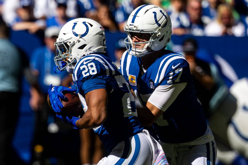 Zwei Footballspieler der Indianapolis Colts in blauen Uniformen und weißen Helmen spielen während eines Spiels. Der Quarterback Daniel Jones scheint den Ball an Jonathan Taylor abzugeben. Eine blau-weiß gekleidete Menschenmenge ist im sonnenbeschienenen Hintergrund verschwommen. Diese Beschreibung wurde mit der FootballR KI automatisch generiert.