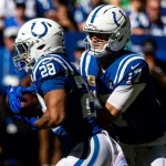 Woche 9 - Zwei Footballspieler der Indianapolis Colts in blauen Uniformen und weißen Helmen spielen während eines Spiels. Der Quarterback Daniel Jones scheint den Ball an Jonathan Taylor abzugeben. Eine blau-weiß gekleidete Menschenmenge ist im sonnenbeschienenen Hintergrund verschwommen. Diese Beschreibung wurde mit der FootballR KI automatisch generiert.