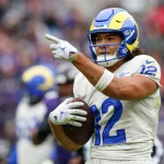 Fantasy Football - NFL Playoff Picture - Puka Nacua, Wide Receiver der Rams, in einer weiß-blauen Uniform und mit Helm, hält einen Football im rechten Arm und zeigt mit der linken Hand. Der Hintergrund ist unscharf und zeigt ein Publikum in einem Stadion. Diese Beschreibung wurde mit der FootballR KI automatisch generiert.